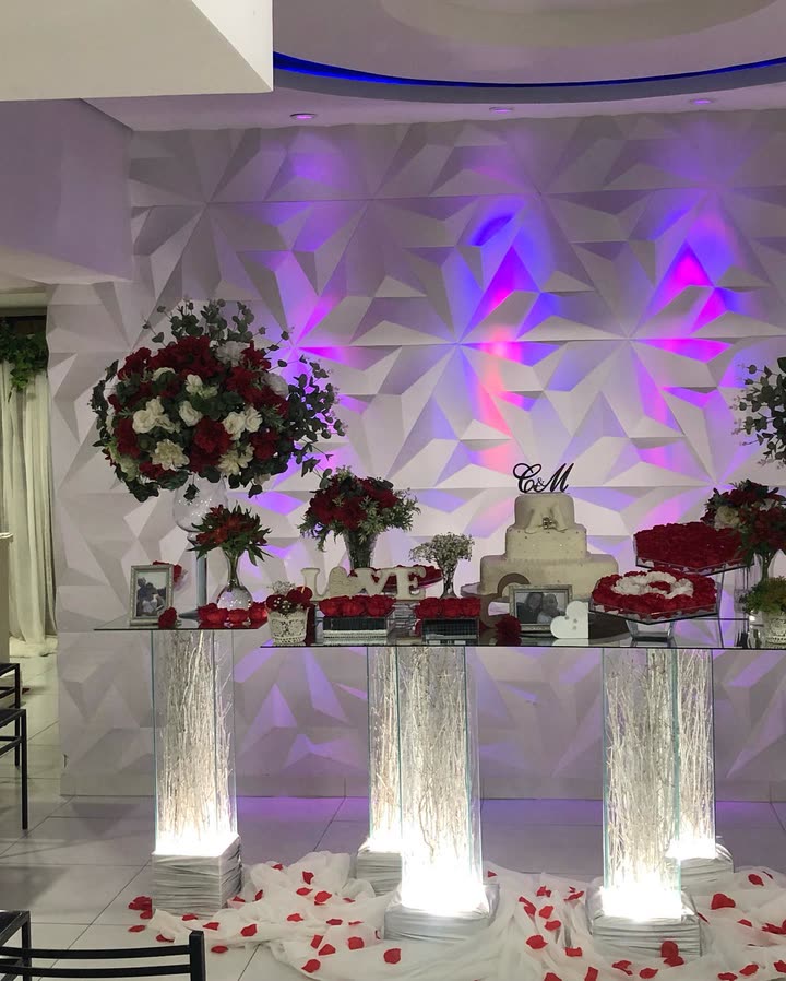Mesa do bolo com arranjos de rosas vermelhas e brancas, iluminada por luzes roxas contra o painel geométrico branco de relevo.