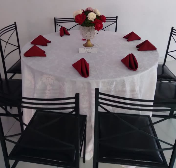 Mesa redonda posta com toalha branca, guardanapos vermelhos dobrados e um arranjo central de flores.