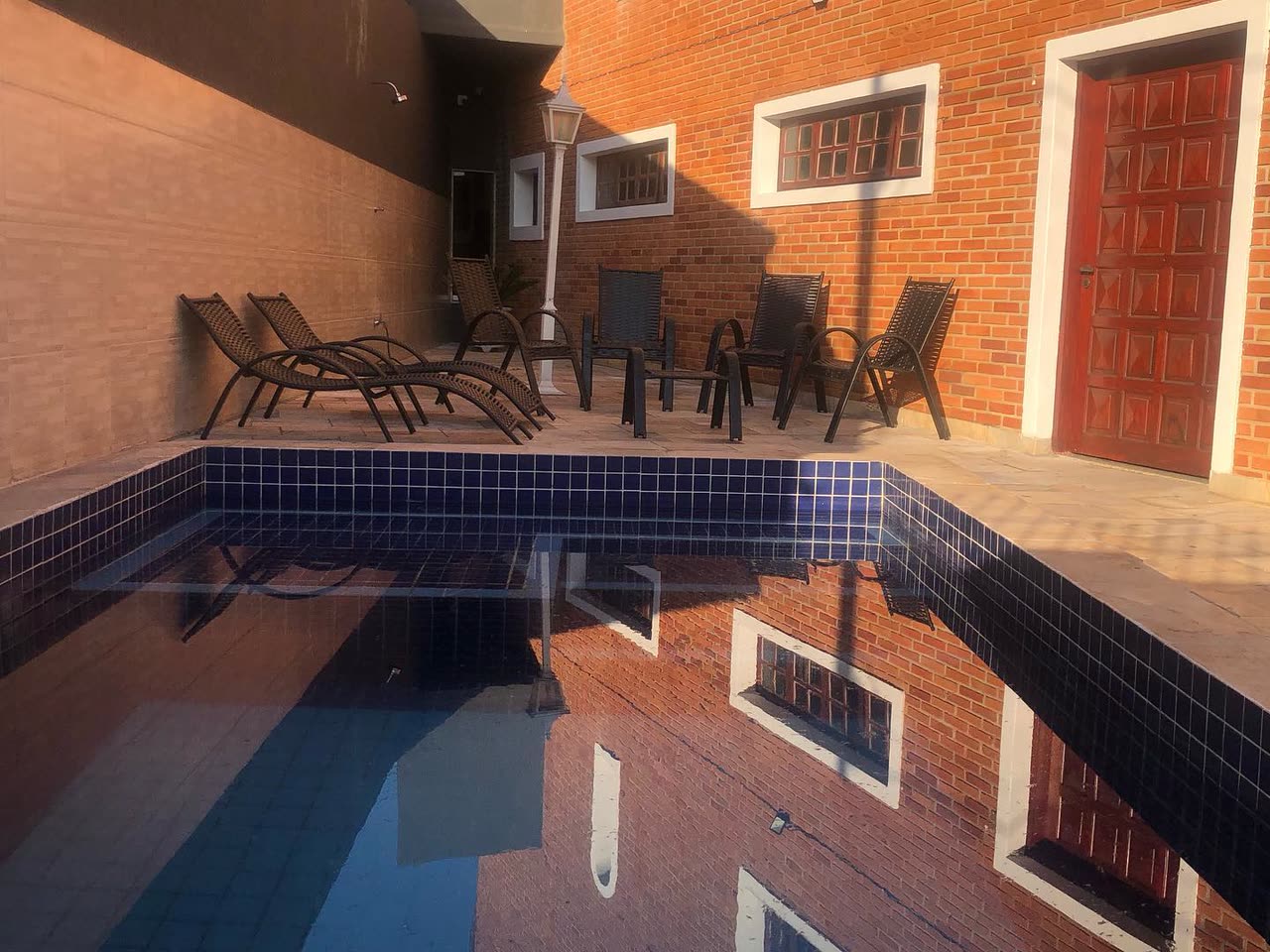 Área externa da Mansão Feest ao fim da tarde: piscina de pastilha azul, espreguiçadeiras e cadeiras de vime junto à parede de tijolo aparente.