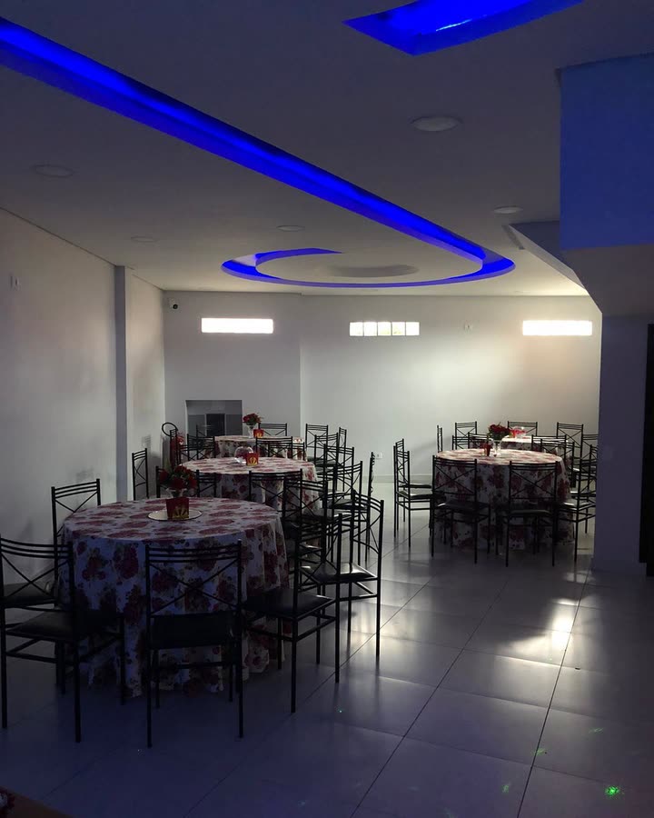 Salão montado com mesas redondas cobertas por toalhas estampadas e cadeiras de ferro pretas, sob a sanca de teto com fita de LED azul.